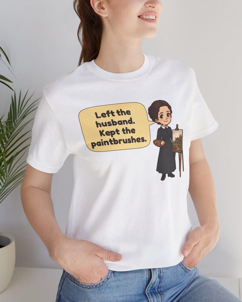 May include: A white short-sleeved t-shirt featuring a cartoon illustration of a person in a black dress standing by an easel with a palette. A yellow speech bubble contains the text: Left the husband. Kept the paintbrushes.