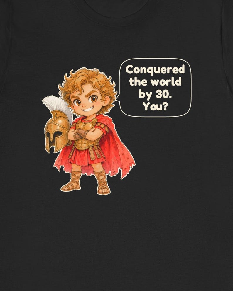 May include: A black t-shirt featuring a cartoon illustration of a young warrior with curly brown hair, wearing gold armor, a red cape, and sandals. The warrior holds a plumed helmet and a speech bubble reads: Conquered the world by 30. You?