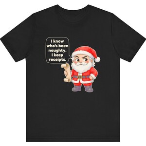 May include: A black t-shirt featuring a cartoon illustration of Santa Claus holding a scroll. Santa wears a red suit with white trim and a black belt. A speech bubble above him contains the text: I know who's been naughty. I keep receipts.