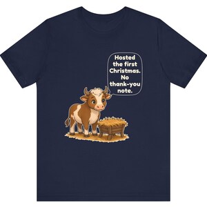 以下が含まれることがあります： ネイビーブルーのTシャツ。干し草が入った木製の飼い葉桶の横に立つ、茶色と白の牛の漫画イラストが描かれています。牛の上の吹き出しには「Hosted the first Christmas. No thank-you note.」というテキストがあります。
