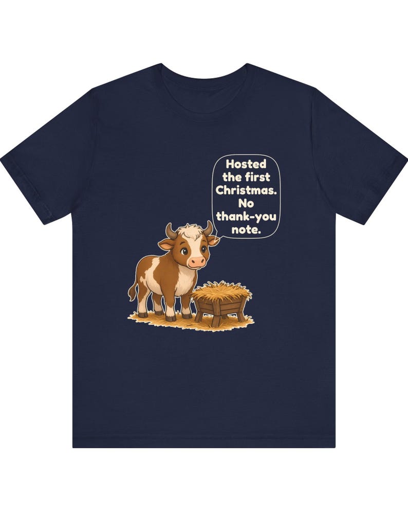 以下が含まれることがあります： ネイビーブルーのTシャツ。干し草が入った木製の飼い葉桶の横に立つ、茶色と白の牛の漫画イラストが描かれています。牛の上の吹き出しには「Hosted the first Christmas. No thank-you note.」というテキストがあります。
