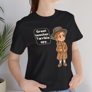 May include: A black short-sleeve t-shirt featuring a cartoon illustration of a person in historical attire wearing a hat and holding a book. A speech bubble next to the character contains the text: Great teacher. Terrible spy.