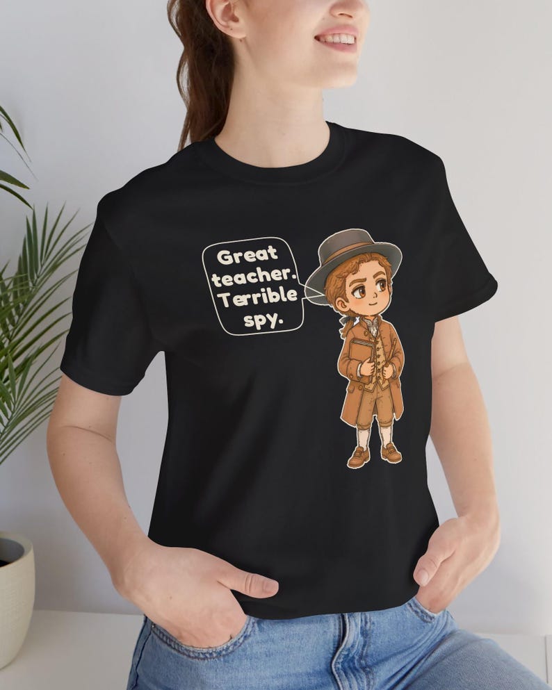 May include: A black short-sleeve t-shirt featuring a cartoon illustration of a person in historical attire wearing a hat and holding a book. A speech bubble next to the character contains the text: Great teacher. Terrible spy.