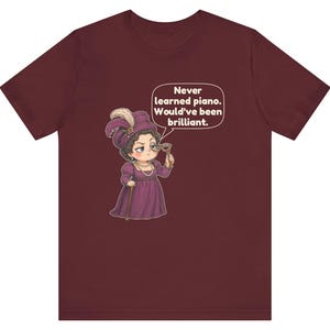 May include: A maroon short-sleeve t-shirt featuring a cartoon illustration of a person in a purple period dress and hat, holding a monocle. A speech bubble contains the text: Never learned piano. Would've been brilliant.