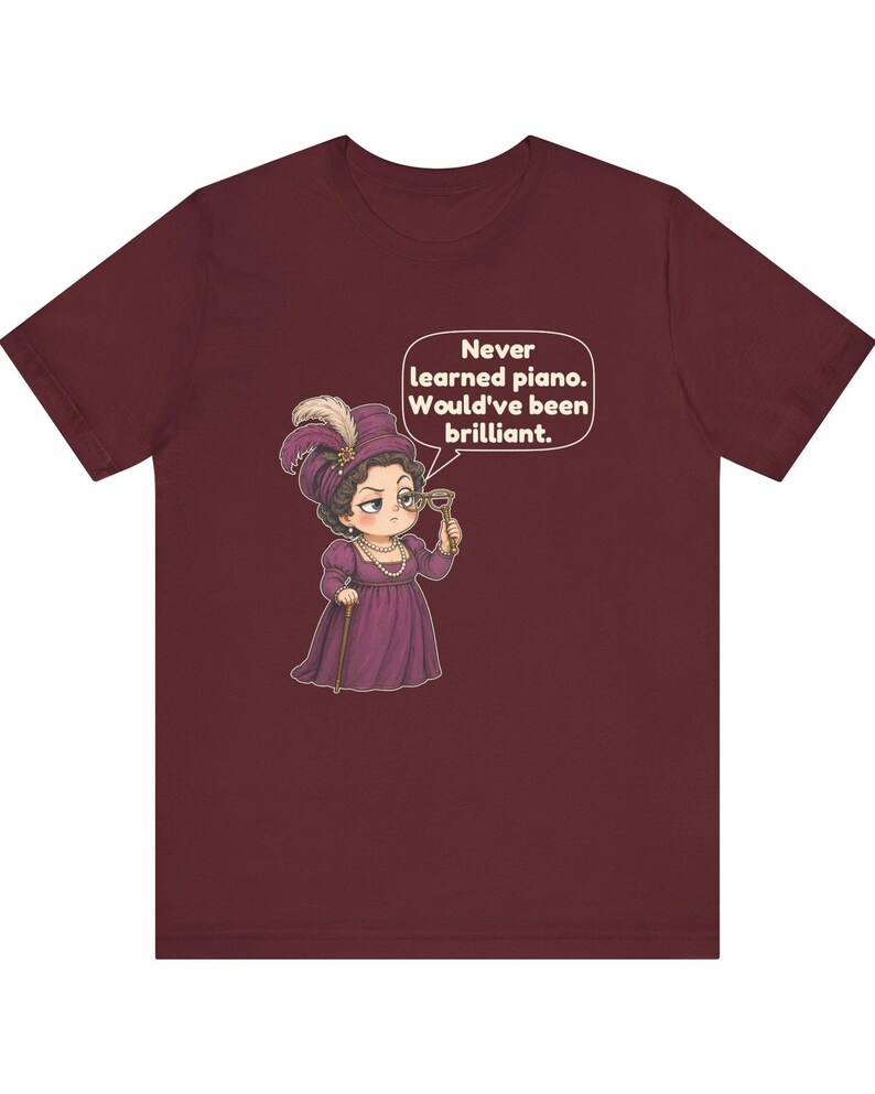 May include: A maroon short-sleeve t-shirt featuring a cartoon illustration of a person in a purple period dress and hat, holding a monocle. A speech bubble contains the text: Never learned piano. Would've been brilliant.