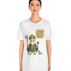 Puede incluir: Una camiseta blanca con una ilustraci&oacute;n de dibujos animados de una figura con vestimentas religiosas verdes que sostiene un bast&oacute;n de madera y un tr&eacute;bol. Una peque&ntilde;a serpiente verde est&aacute; cerca. Un globo de texto contiene: I drove out the snakes. You're next, Monday.