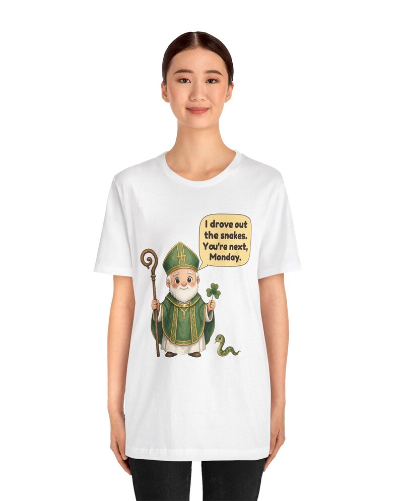 Puede incluir: Una camiseta blanca con una ilustraci&oacute;n de dibujos animados de una figura con vestimentas religiosas verdes que sostiene un bast&oacute;n de madera y un tr&eacute;bol. Una peque&ntilde;a serpiente verde est&aacute; cerca. Un globo de texto contiene: I drove out the snakes. You're next, Monday.