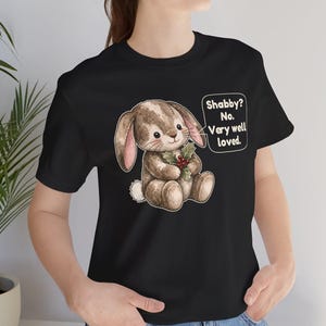 May include: A black t-shirt featuring a graphic of a brown stuffed rabbit holding a sprig of holly. A speech bubble next to the rabbit contains the text: Shabby? No. Very well loved. The rabbit has stitched details and a white tail.