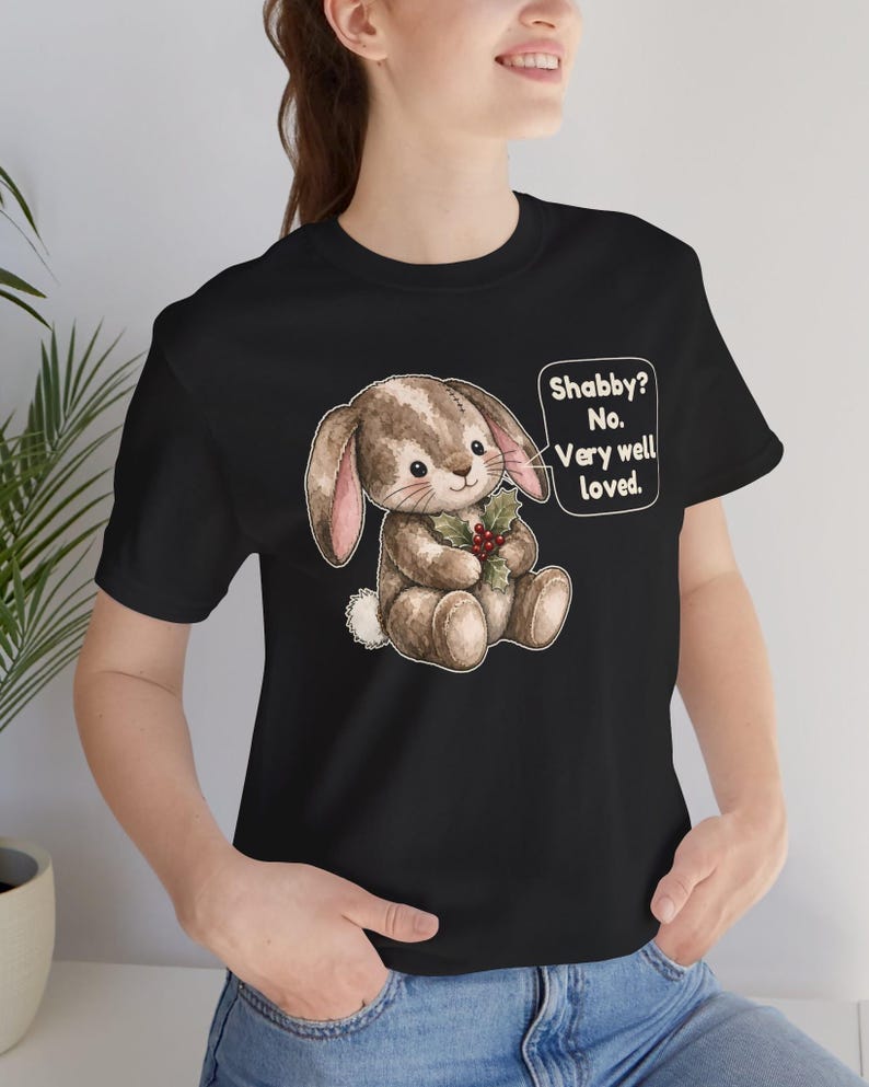 May include: A black t-shirt featuring a graphic of a brown stuffed rabbit holding a sprig of holly. A speech bubble next to the rabbit contains the text: Shabby? No. Very well loved. The rabbit has stitched details and a white tail.