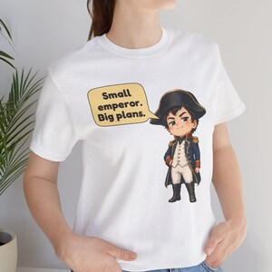 May include: A white short-sleeved t-shirt featuring a cartoon illustration of a historical figure in a blue military uniform and black hat. A yellow speech bubble next to the character contains the text: Small emperor. Big plans.