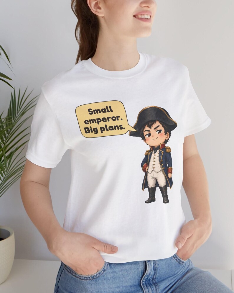 May include: A white short-sleeved t-shirt featuring a cartoon illustration of a historical figure in a blue military uniform and black hat. A yellow speech bubble next to the character contains the text: Small emperor. Big plans.