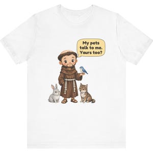 May include: A white t-shirt featuring a cartoon illustration of a monk in a brown robe standing with a grey rabbit, a brown wolf, and a blue bird. A speech bubble above the monk contains the text: My pets talk to me. Yours too?