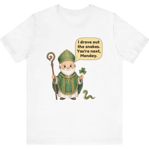Puede incluir: Una camiseta blanca con una ilustraci&oacute;n de dibujos animados de una figura con vestiduras religiosas verdes que sostiene un bast&oacute;n de madera y un tr&eacute;bol. Un globo de texto contiene: I drove out the snakes. You're next, Monday. Una peque&ntilde;a serpiente verde est&aacute; en el suelo.