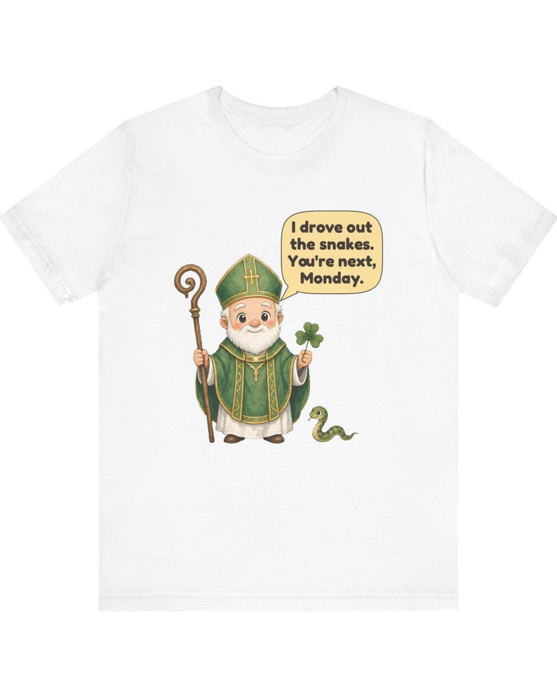 Puede incluir: Una camiseta blanca con una ilustraci&oacute;n de dibujos animados de una figura con vestiduras religiosas verdes que sostiene un bast&oacute;n de madera y un tr&eacute;bol. Un globo de texto contiene: I drove out the snakes. You're next, Monday. Una peque&ntilde;a serpiente verde est&aacute; en el suelo.