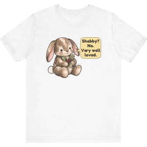 May include: A white short-sleeve t-shirt featuring a graphic of a brown bunny holding a sprig of holly. Next to the bunny is a yellow speech bubble containing the text: Shabby? No. Very well loved. The design is centered on the chest area.