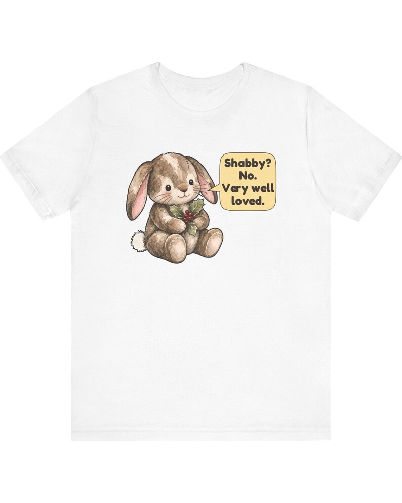 May include: A white short-sleeve t-shirt featuring a graphic of a brown bunny holding a sprig of holly. Next to the bunny is a yellow speech bubble containing the text: Shabby? No. Very well loved. The design is centered on the chest area.