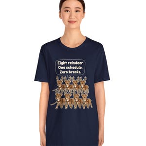 K&ouml;nnte beinhalten: Ein dunkelblaues Kurzarm-T-Shirt mit einer Grafik von acht braunen Rentieren in zwei Reihen. &Uuml;ber den Rentieren befindet sich eine Sprechblase mit dem wei&szlig;en Text: Eight reindeer. One schedule. Zero breaks.