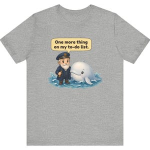 May include: A heather gray t-shirt featuring a graphic of a cartoon sea captain with a white beard and a white whale in blue ocean waves. A yellow speech bubble above them contains the black text, One more thing on my to-do list.