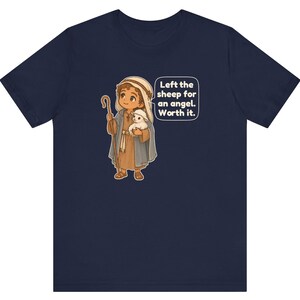 May include: A navy blue t-shirt featuring a cartoon illustration of a shepherd holding a small white lamb and a wooden staff. A speech bubble next to the character contains the text: Left the sheep for an angel. Worth it.