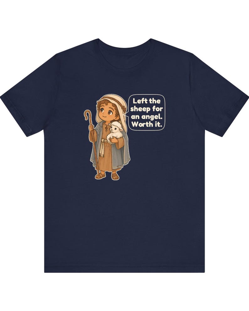 May include: A navy blue t-shirt featuring a cartoon illustration of a shepherd holding a small white lamb and a wooden staff. A speech bubble next to the character contains the text: Left the sheep for an angel. Worth it.