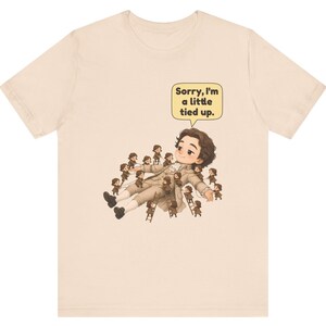 Op de afbeelding: Een beige t-shirt met korte mouwen en een afbeelding van een groot persoon die ligt, omringd door vele kleine mensen die touwen en ladders gebruiken om hen vast te binden. Een tekstballon bevat de tekst, Sorry, I'm a little tied up.