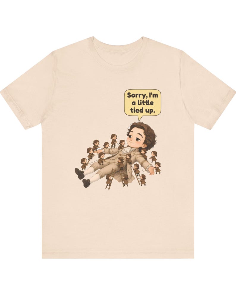 Op de afbeelding: Een beige t-shirt met korte mouwen en een afbeelding van een groot persoon die ligt, omringd door vele kleine mensen die touwen en ladders gebruiken om hen vast te binden. Een tekstballon bevat de tekst, Sorry, I'm a little tied up.