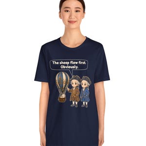 May include: A navy blue short-sleeve t-shirt featuring a graphic of two historical figures in 18th-century attire standing next to a hot air balloon with a sheep inside. Above the figures, a speech bubble contains the text: The sheep flew first. Obviously.