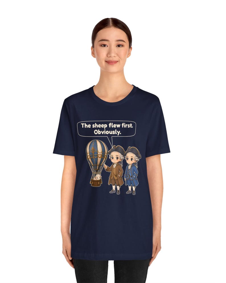 May include: A navy blue short-sleeve t-shirt featuring a graphic of two historical figures in 18th-century attire standing next to a hot air balloon with a sheep inside. Above the figures, a speech bubble contains the text: The sheep flew first. Obviously.
