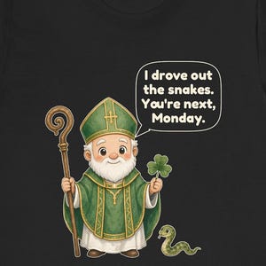 Puede incluir: Una camiseta negra con una ilustraci&oacute;n de dibujos animados de una figura barbuda con vestiduras religiosas verdes y una mitra, sosteniendo un bast&oacute;n de madera y un tr&eacute;bol. Una peque&ntilde;a serpiente verde est&aacute; cerca. Un bocadillo contiene el texto: I drove out the snakes. You're next, Monday.