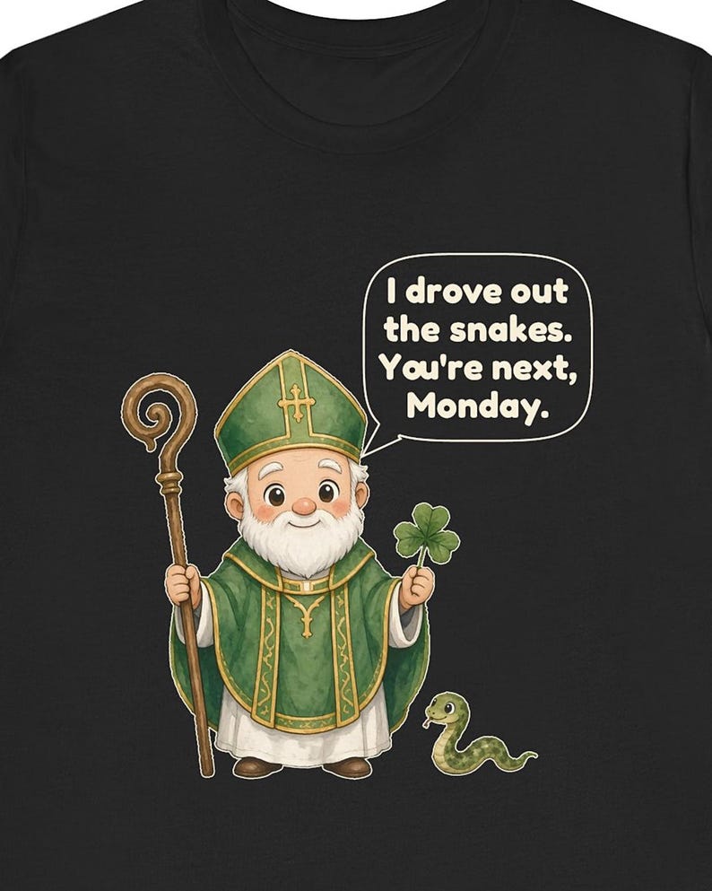 Puede incluir: Una camiseta negra con una ilustraci&oacute;n de dibujos animados de una figura barbuda con vestiduras religiosas verdes y una mitra, sosteniendo un bast&oacute;n de madera y un tr&eacute;bol. Una peque&ntilde;a serpiente verde est&aacute; cerca. Un bocadillo contiene el texto: I drove out the snakes. You're next, Monday.