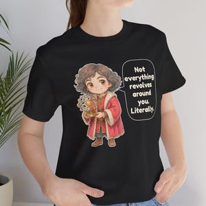 May include: A black t-shirt featuring a cartoon illustration of a person with curly brown hair wearing a red coat with fur trim, holding a golden armillary sphere. A speech bubble contains the text: Not everything revolves around you. Literally.