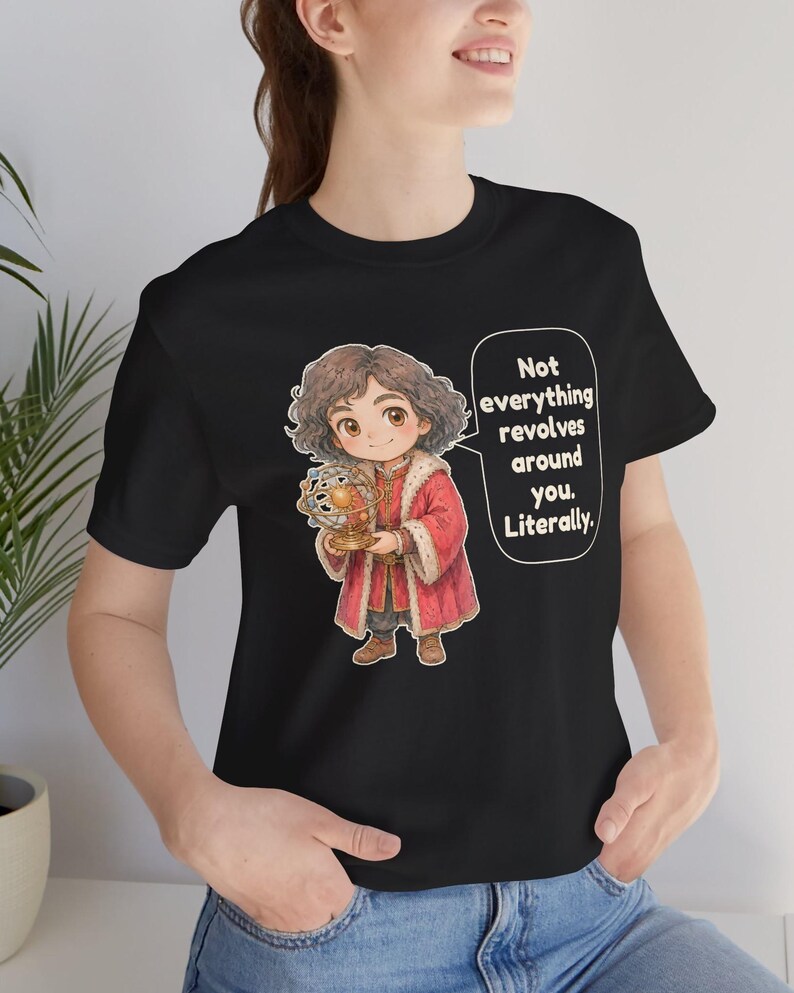 May include: A black t-shirt featuring a cartoon illustration of a person with curly brown hair wearing a red coat with fur trim, holding a golden armillary sphere. A speech bubble contains the text: Not everything revolves around you. Literally.
