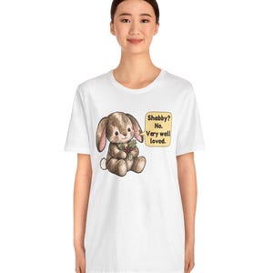 May include: A white t-shirt featuring a brown bunny illustration holding a sprig of holly. Next to the bunny is a yellow speech bubble with the text: Shabby? No. Very well loved. The design is centered on the chest of the garment.