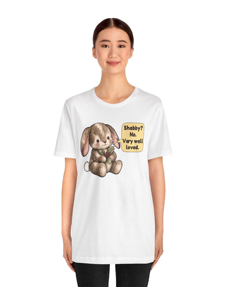 May include: A white t-shirt featuring a brown bunny illustration holding a sprig of holly. Next to the bunny is a yellow speech bubble with the text: Shabby? No. Very well loved. The design is centered on the chest of the garment.