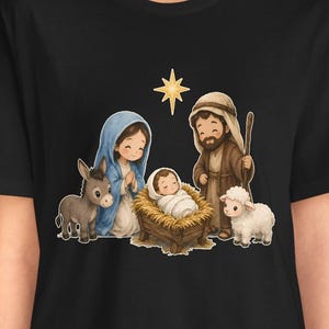 May include: A black t-shirt featuring a cartoon-style nativity scene graphic. The illustration depicts a person in a blue head covering, a person in a brown robe with a staff, a baby in a manger, a donkey, a sheep, and a yellow star.