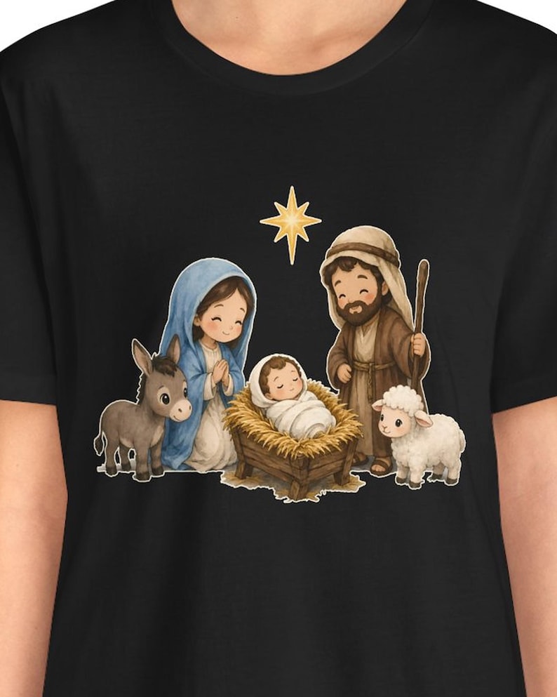 May include: A black t-shirt featuring a cartoon-style nativity scene graphic. The illustration depicts a person in a blue head covering, a person in a brown robe with a staff, a baby in a manger, a donkey, a sheep, and a yellow star.
