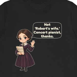 May include: A black t-shirt featuring a cartoon illustration of a person in a dark purple dress holding sheet music. A speech bubble contains the text: Not 'Robert's wife.' Concert pianist, thanks.