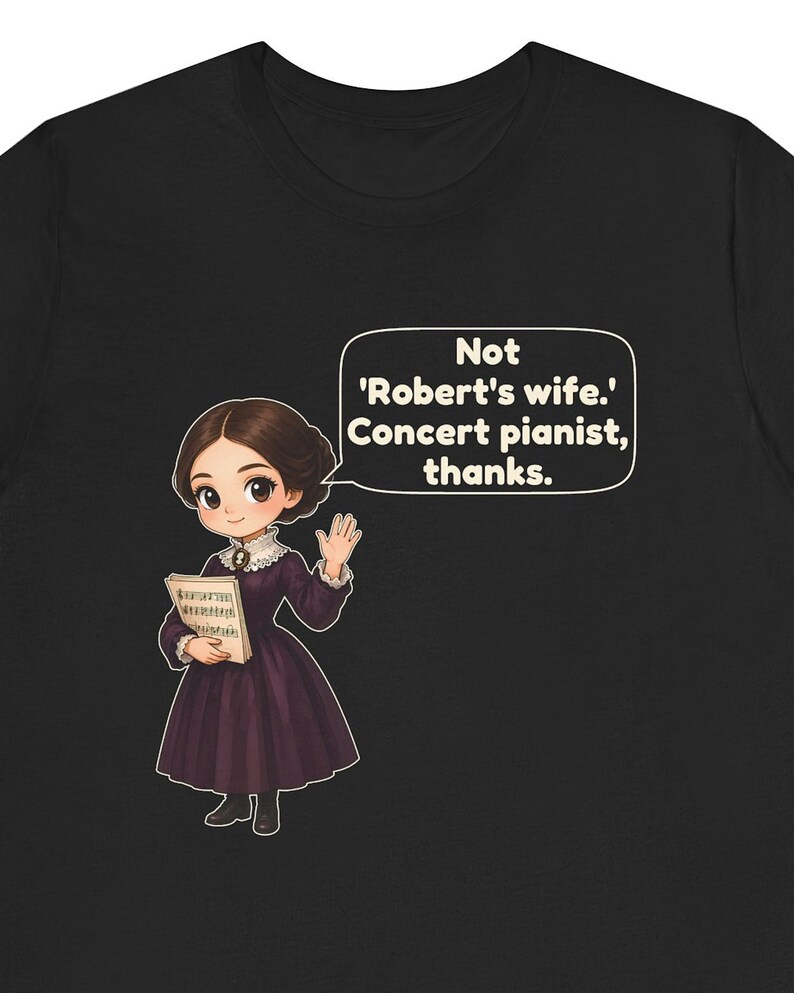 Puede incluir: Una camiseta negra con una ilustraci&oacute;n de dibujos animados de una persona con un vestido morado oscuro que sostiene partituras. Un bocadillo contiene el texto: Not 'Robert's wife.' Concert pianist, thanks.