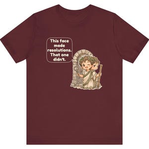 K&ouml;nnte beinhalten: Ein kastanienbraunes T-Shirt mit einer Grafik einer zweigesichtigen Figur, die einen Lorbeerkranz und eine Toga tr&auml;gt und vor einem Steinbogen steht. Die Figur h&auml;lt einen Schl&uuml;ssel und einen Stab. Eine Sprechblase enth&auml;lt den Text: This face made resolutions. That one didn't.