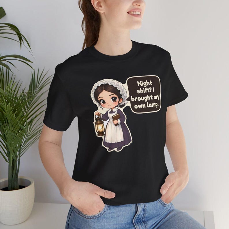 Pode incluir: Uma t-shirt preta de manga curta com uma ilustra&ccedil;&atilde;o de desenho animado de uma enfermeira com touca branca e vestido roxo segurando uma lanterna e um livro. Um bal&atilde;o de fala cont&eacute;m o texto: Night shift? I brought my own lamp.