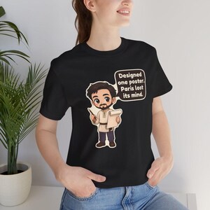 May include: A black short-sleeve t-shirt featuring a cartoon illustration of a bearded person holding a pencil and a scroll. A speech bubble next to the character contains the text: Designed one poster. Paris lost its mind.