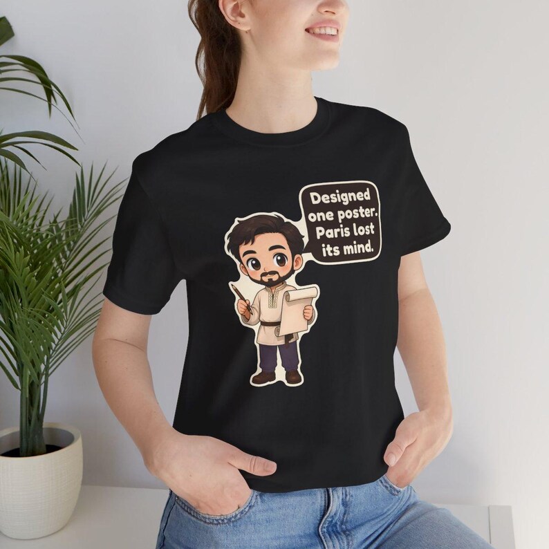 May include: A black short-sleeve t-shirt featuring a cartoon illustration of a bearded person holding a pencil and a scroll. A speech bubble next to the character contains the text: Designed one poster. Paris lost its mind.