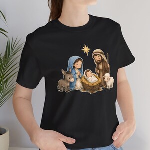 May include: A black t-shirt featuring a cartoon-style nativity scene graphic. The illustration depicts a figure in a blue head covering, a figure in brown robes, a baby in a manger, a donkey, a lamb, and a yellow star above them.