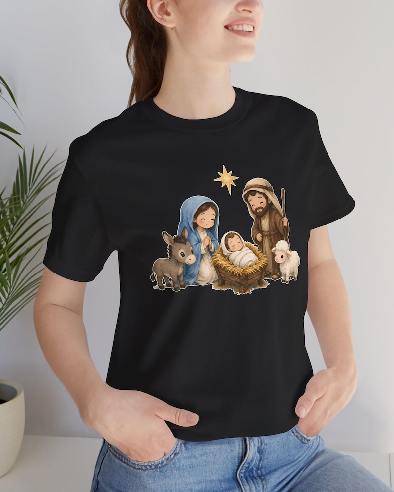 May include: A black t-shirt featuring a cartoon-style nativity scene graphic. The illustration depicts a figure in a blue head covering, a figure in brown robes, a baby in a manger, a donkey, a lamb, and a yellow star above them.