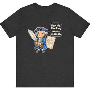 May include: A dark heather gray t-shirt featuring a cartoon illustration of a person in 18th-century blue attire holding a white quill and a scroll. A speech bubble contains the text: Sign big. The king needs glasses.