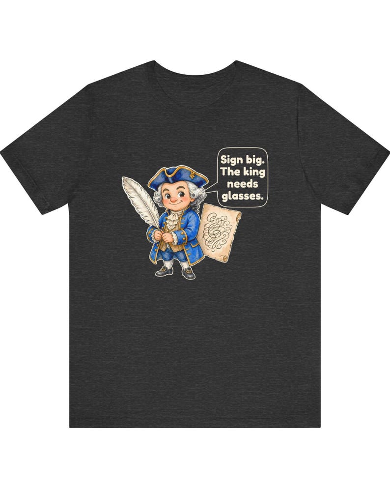 May include: A dark heather gray t-shirt featuring a cartoon illustration of a person in 18th-century blue attire holding a white quill and a scroll. A speech bubble contains the text: Sign big. The king needs glasses.