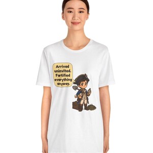 May include: A white t-shirt featuring a cartoon illustration of a historical figure in a blue coat and tricorn hat holding a shovel and compass, with a speech bubble that reads: Arrived uninvited. Fortified everything anyway.