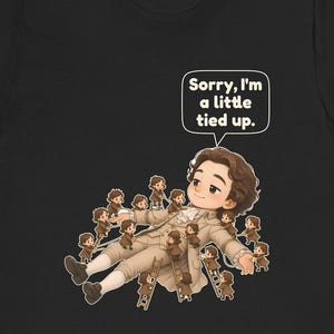 Op de afbeelding: Een zwart t-shirt met een afbeelding van een groot persoon in 18e-eeuwse kleding die ligt, omringd door vele kleine figuren die touwen en ladders gebruiken om hen vast te binden. Een tekstballon boven de grote figuur bevat de tekst, Sorry, I'm a little tied up.