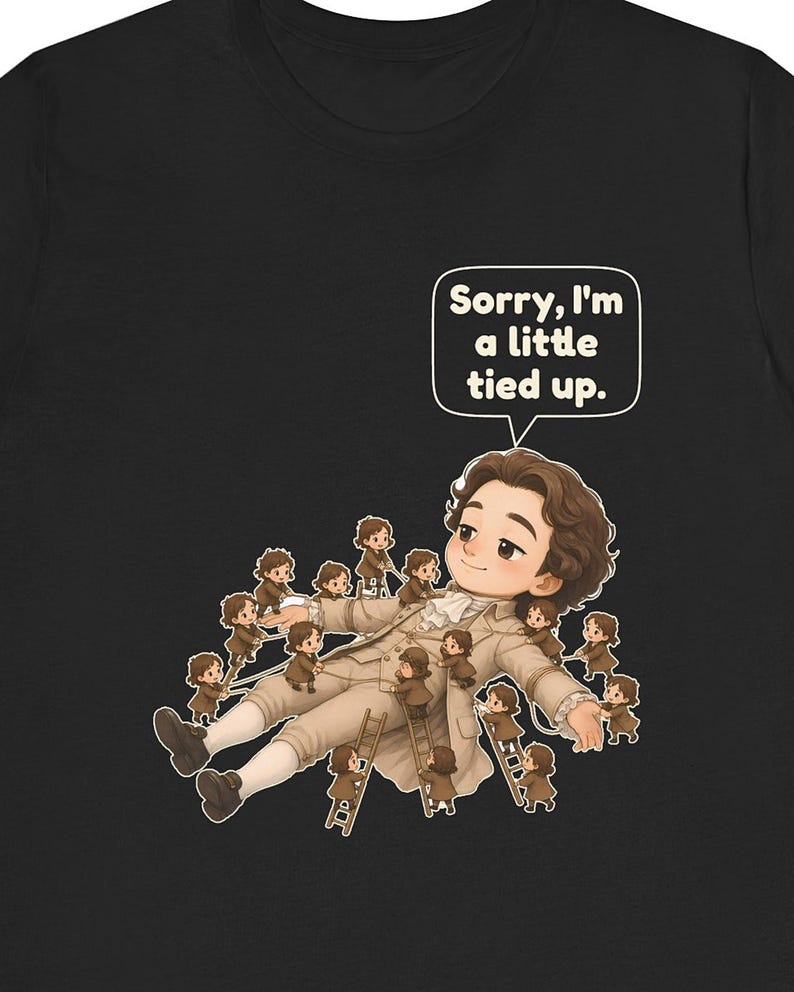 Op de afbeelding: Een zwart t-shirt met een afbeelding van een groot persoon in 18e-eeuwse kleding die ligt, omringd door vele kleine figuren die touwen en ladders gebruiken om hen vast te binden. Een tekstballon boven de grote figuur bevat de tekst, Sorry, I'm a little tied up.
