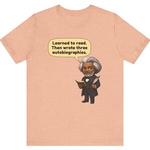 Puede incluir: Una camiseta de color melocot&oacute;n con una ilustraci&oacute;n de dibujos animados de Frederick Douglass sosteniendo un libro y una pluma. Un globo de texto sobre la figura contiene el texto: Learned to read. Then wrote three autobiographies.